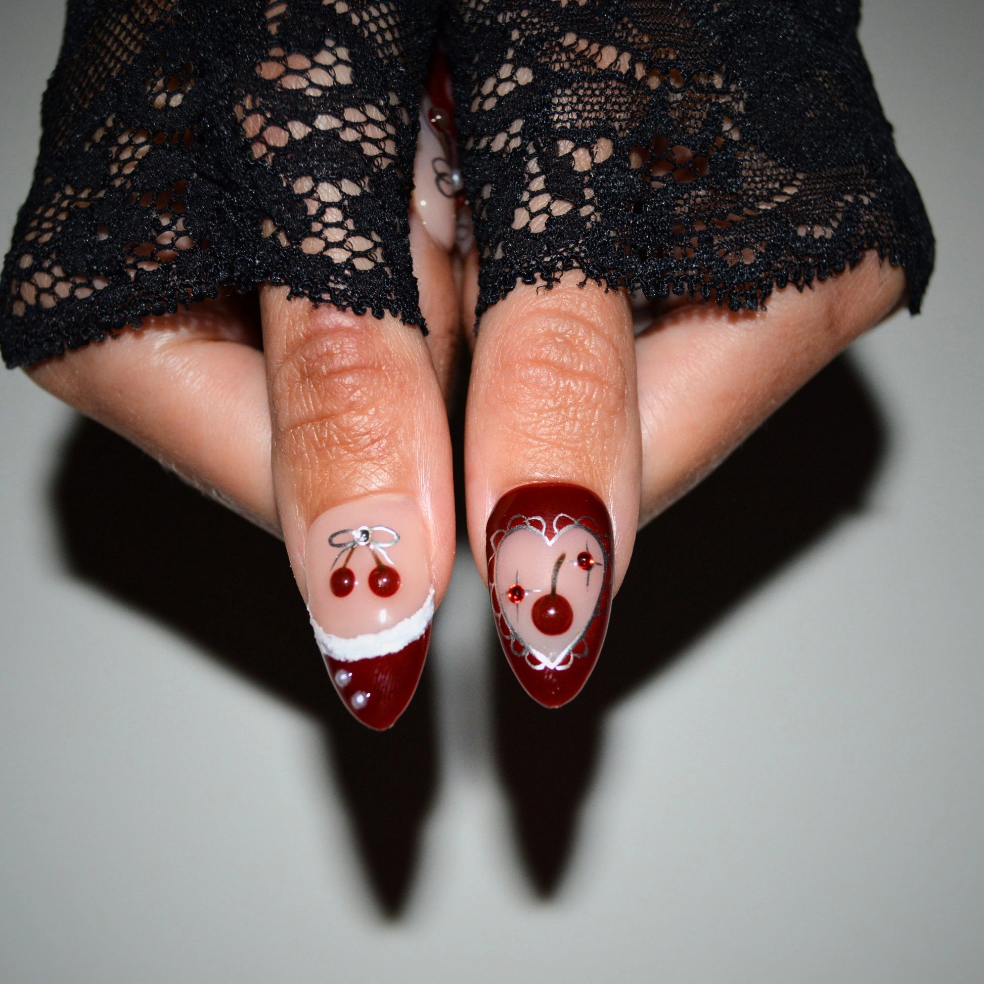 Gros plan sur deux ongles press-on Cherry Christmas faits main en gel et acrylique haut de gamme, décorés d’une cerise 3D et d’un cœur rouge bordé d’argent.