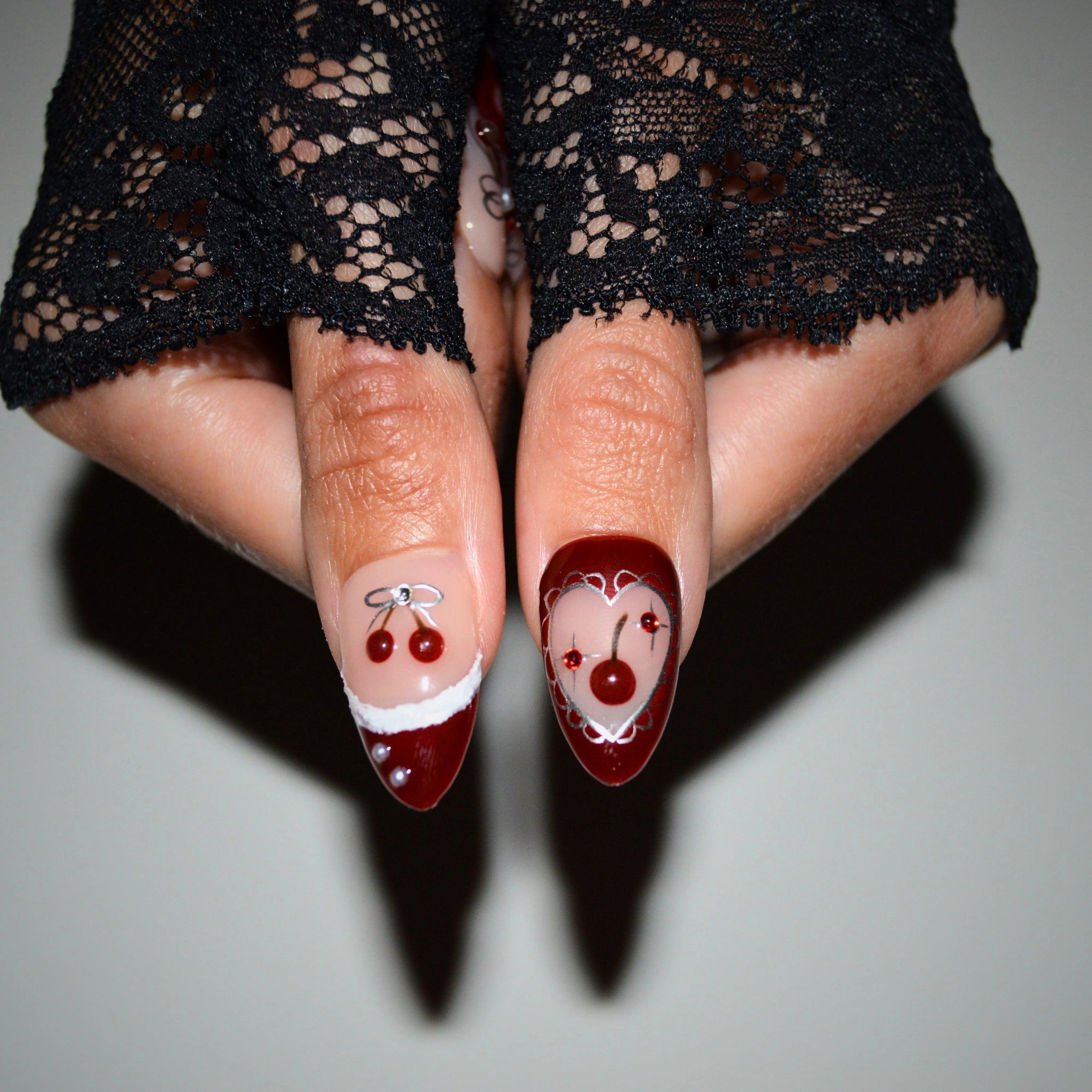 Gros plan sur deux ongles press-on Cherry Christmas faits main en gel et acrylique haut de gamme, décorés d’une cerise 3D et d’un cœur rouge bordé d’argent.