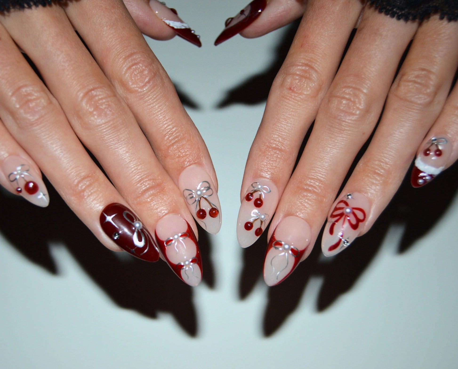 Press-on nails Cherry Christmas faits main en gel et acrylique, design nude et rouge avec cerises 3D, nœuds métalliques et finitions festives.