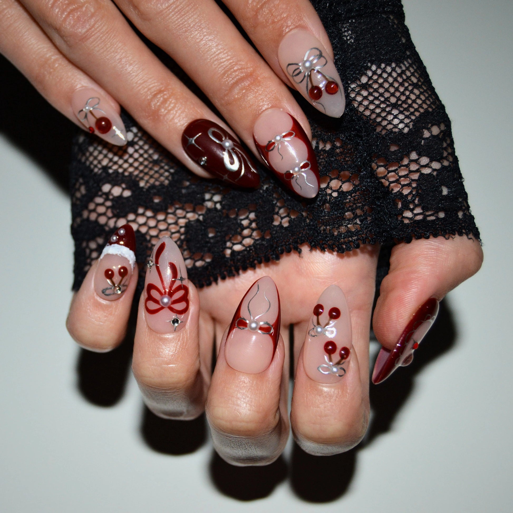 Ongles press-on Cherry Christmas faits main en gel et acrylique haut de gamme, décorés de cerises rouges, nœuds argentés et détails de Noël sur forme amande.