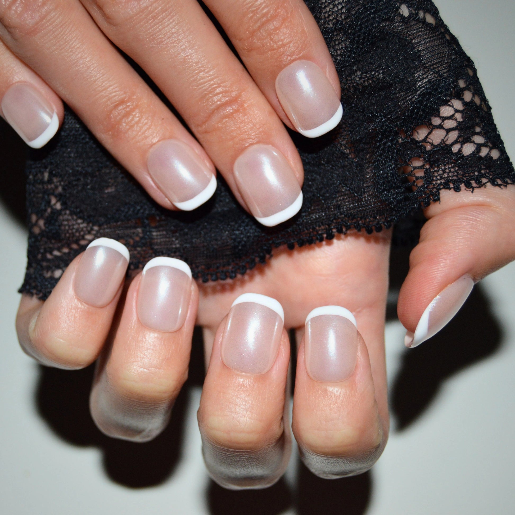 Press-on nails carrés French courts, gel & acrylique, vue rapprochée main tenant dentelle noire.